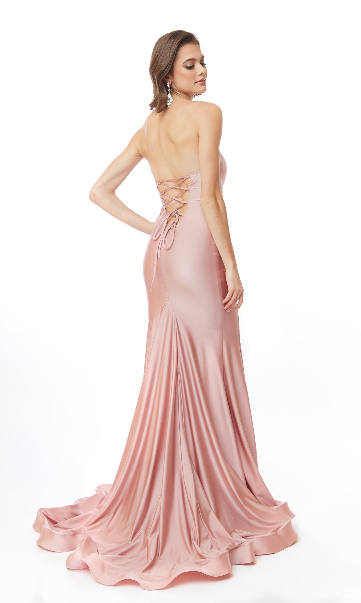 A woman stands against a white background, looking over her shoulder in the Atria 6544H Lace-Up Strapless Long Prom Dress—a floor-length pink satin gown with a lace-up back.